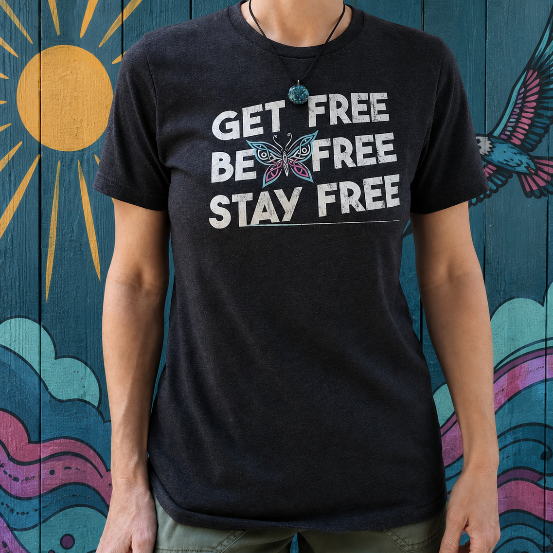 A person wearing a dark charcoal heather t-shirt with a graphic that reads 'GET FREE BE FREE STAY FREE' in white text, featuring a butterfly design. The background is a painted wooden wall with a yellow sun, blue bird, and colorful waves.