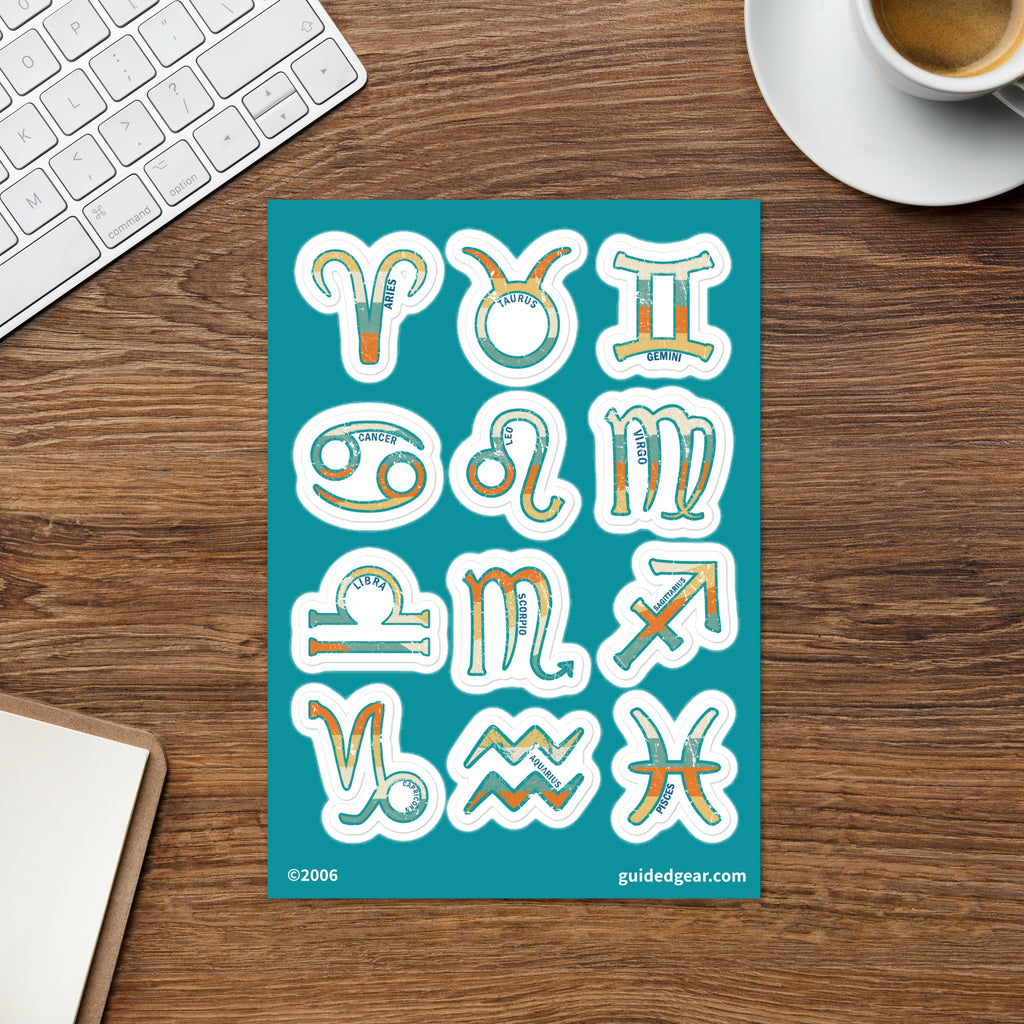 A teal sticker sheet displaying twelve zodiac symbols in a retro, hand-drawn style with orange and yellow accents, styled on a wooden desk next to a keyboard, coffee cup, and notebook.