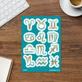 A teal sticker sheet displaying twelve zodiac symbols in a retro, hand-drawn style with orange and yellow accents, styled on a wooden desk next to a keyboard, coffee cup, and notebook.