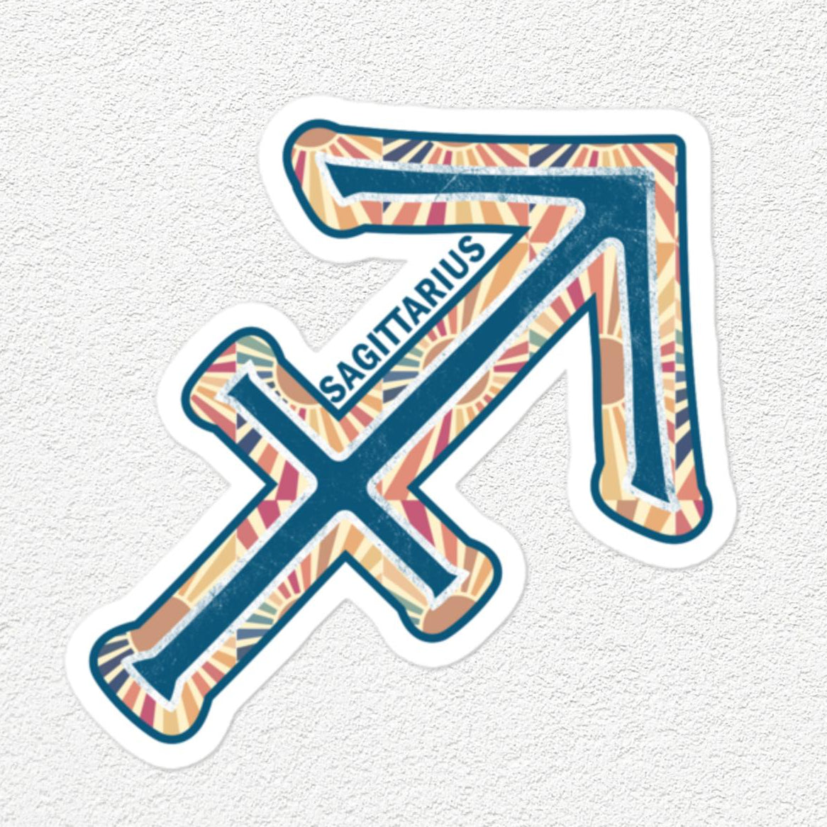 A die-cut sticker featuring the Sagittarius zodiac symbol with a colorful, patterned fill and the word 'SAGITTARIUS' printed along the arrow, shown on a textured off-white background.