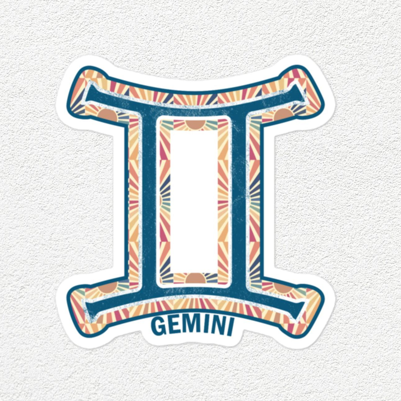 A die-cut vinyl sticker of the Gemini zodiac symbol, featuring a colorful geometric pattern in orange, pink, and blue on a white background with the word 'GEMINI' printed below, shown on a light textured wall.