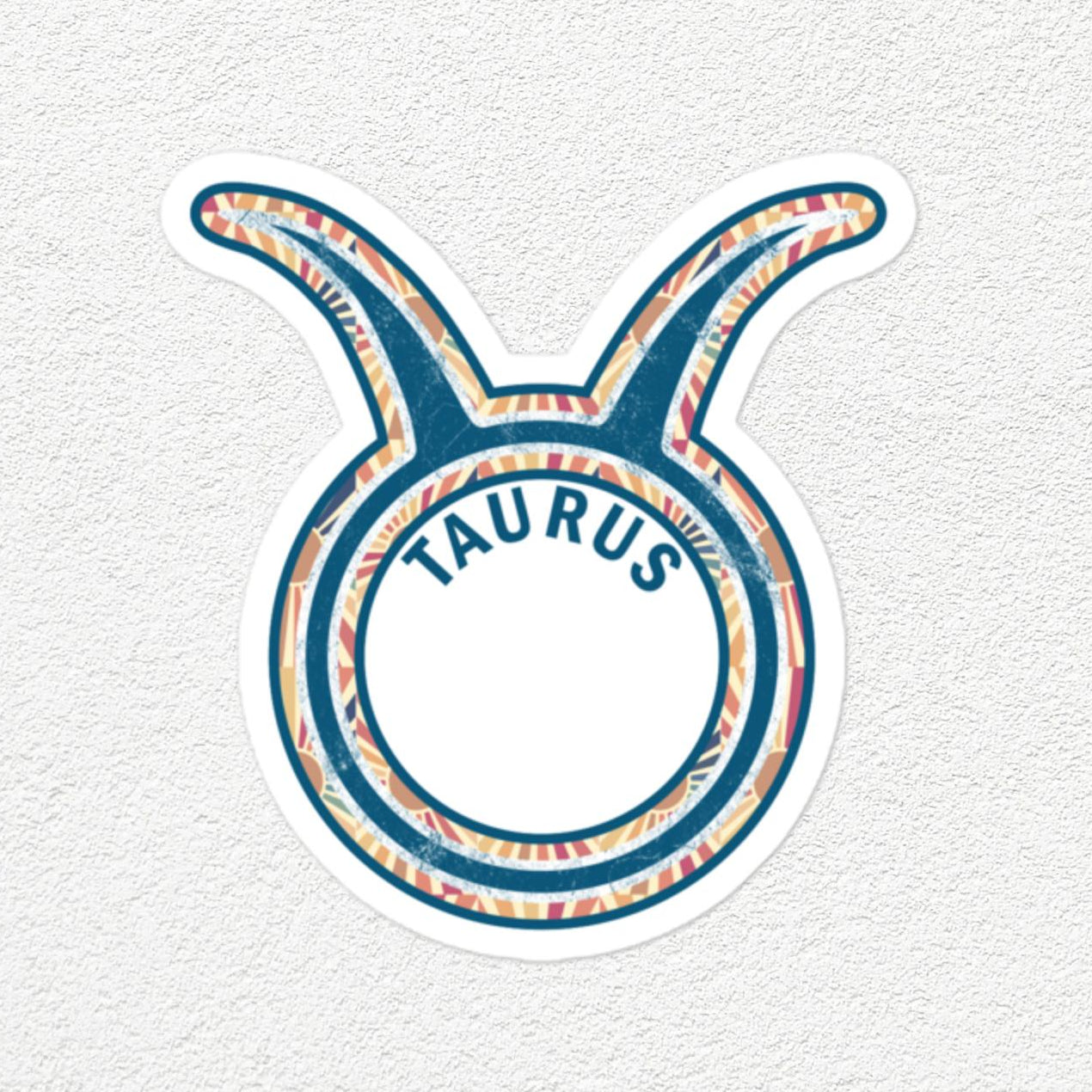 A die-cut vinyl sticker of the Taurus zodiac sign, featuring a blue and white patterned design with the word 'TAURUS' in blue text on a white circular background, set against a textured off-white wall.