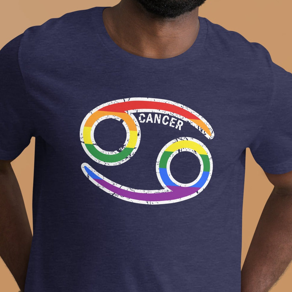 A smiling man wearing a navy blue t-shirt with a colorful rainbow Cancer zodiac symbol printed on the chest, set against a solid warm brown background.