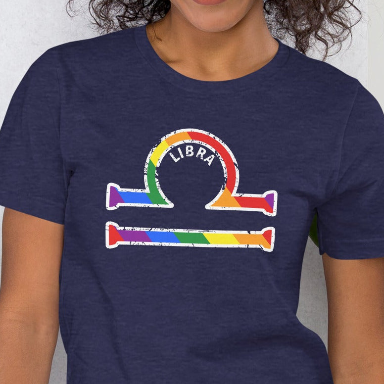Woman wearing a t-shirt with a rainbow Libra astrology symbol graphic design, standing against a white wall.