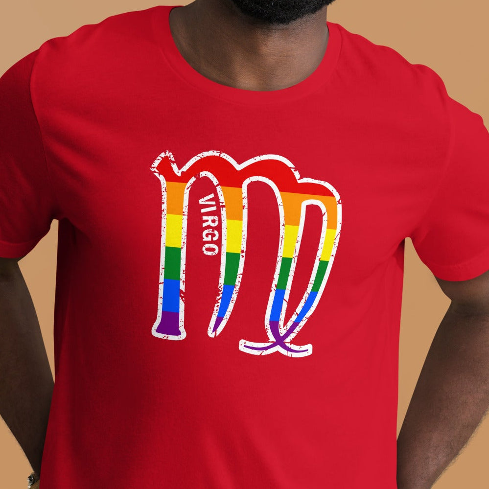 A smiling man wearing a bright red t-shirt with a rainbow-striped Virgo zodiac symbol printed on the chest, set against a solid warm tan background.