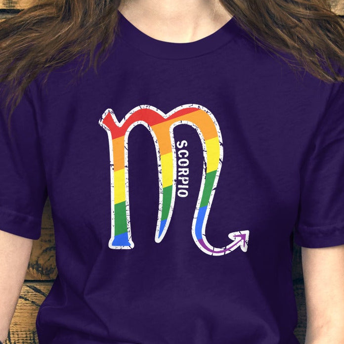 A woman wearing a purple t-shirt with a rainbow-colored 'Scorpio' graphic and the word 'Scorpio' printed on the front, sitting against a rustic wooden wall next to a red and white helmet.