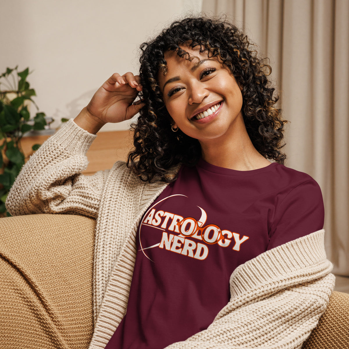 A smiling woman with curly hair sits on a beige couch wearing a burgundy graphic T-shirt with 'ASTROLOGY NERD' printed in white and orange, layered under a cream knit cardigan.